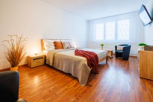 Světlý dvoulůžkový pokoj s béžovou přikrývkou, červeným přehozem a modrým křeslem