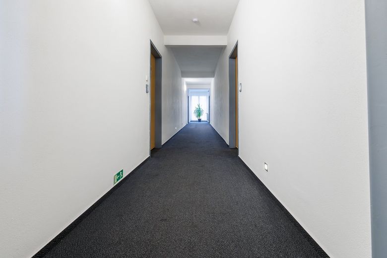Chodba hotelu s tmavě šedým kobercem, dveřmi pokojů a oknem na konci